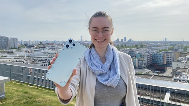Eine Person mit Brille, hellblau gemustertem Schal, grauem T-Shirt und beiger Jacke steht auf einer Dachterrasse und hält ein Smartphone mit heller Rückseite und drei Kameralinsen in die Kamera. Im Hintergrund ist eine weitläufige Stadtlandschaft mit Wohngebäuden, Hochhäusern und vereinzelten Kirchtürmen zu sehen. Der Himmel ist leicht bewölkt, aber hell.