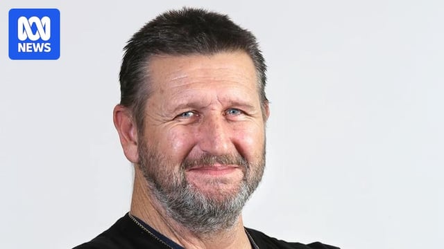 white man with greying hair and beard in chain and black top with arm tattoos smiles at camera with arms crossed