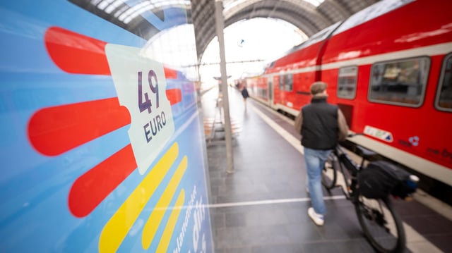 Ein Mann geht an einem Bahnhof mit seinem Fahrrad zwischen der Werbung für das Deutschlandticket und einer Regionalbahn entlang. 