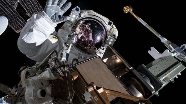 A NASA astronaut conducts a spacewalk outside the International Space Station, with the reflection of Earth and station hardware visible on the helmet visor. The astronaut is surrounded by solar panel structures and robotic tools, captured mid-mission in Earth’s orbit.
