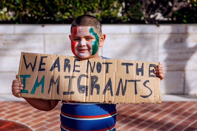 A lttle boy in red, white and green face paint holds a handwritten sign reading "WE AEW NOT THE IMMIGRANTS" as he participates in an outdoor protest
