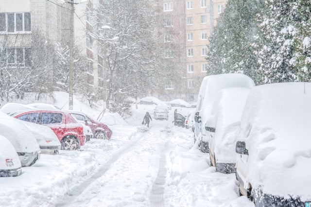 A heavy snowstorm blankets a city street with parked cars as a lone person walks down the avenue.