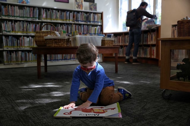 Frank, 2, opens flaps while reading a book as his father, Shawn, looks for books at the Oakland Public Library Rockridge Branch in 2021.