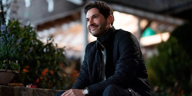 Tom Ellis as Oliver sitting down while looking up and smiling in Season 2 of Tell Me Lies