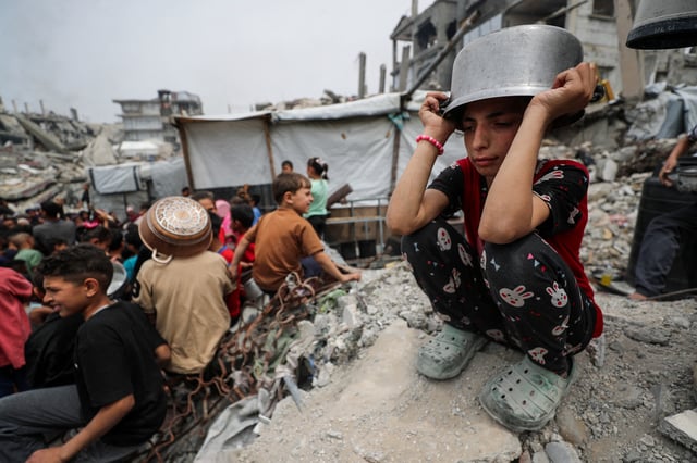 A girl puts a pot to her head as Palestinians wait to receive food cooked by a charity kitchen, in Beit Lahia, northern Gaza Strip, April 24, 2025. REUTERS/Mahmoud Issa/File Photo