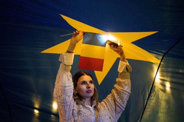 A woman holding a Romanian flag shines the light of her mobile phone as she stands under a large European Union flag during a pro-EU rally ahead of the second round of the presidential election redo in Bucharest, Romania, Friday, May 9, 2025. (AP Photo/Andreea Alexandru)