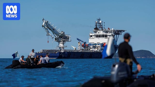 Two small boats with people in uniform in the foreground and a large ship with a crane on it in the background.