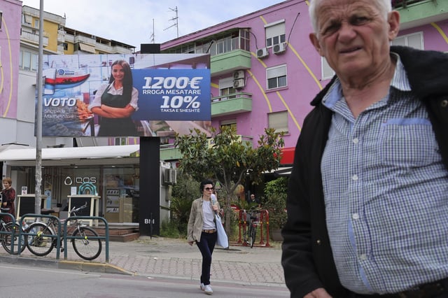 People pass a billboard of the opposition Democratic Party (ASHM) that reads "Vote Grandiose Albania, 1,200 euros average salary, 10% flat tax," in Tirana, Albania, Friday, May 9, 2025. (AP Photo/Vlasov Sulaj)
