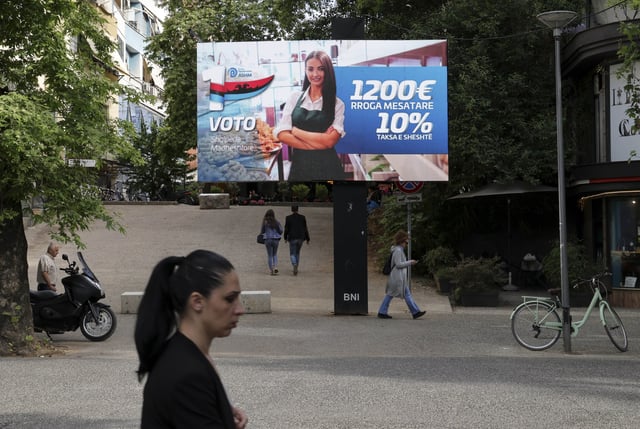 People pass a billboard of the opposition Democratic Party (ASHM) that reads "Vote Grandiose Albania, 1,200 euros average salary, 10% flat tax," in Tirana, Albania, Friday, May 9, 2025. (AP Photo/Vlasov Sulaj)