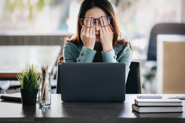 A stock image of a woman sitting at a work desk with her hands over her eyes under her glasses.