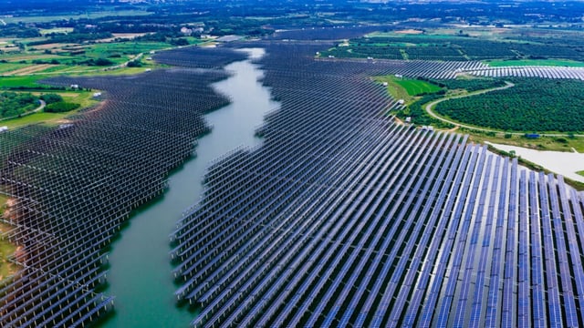 A large solar installation, with panels encroaching on a body of water in the center from both banks. Many panels are installed on stanchions in the water.