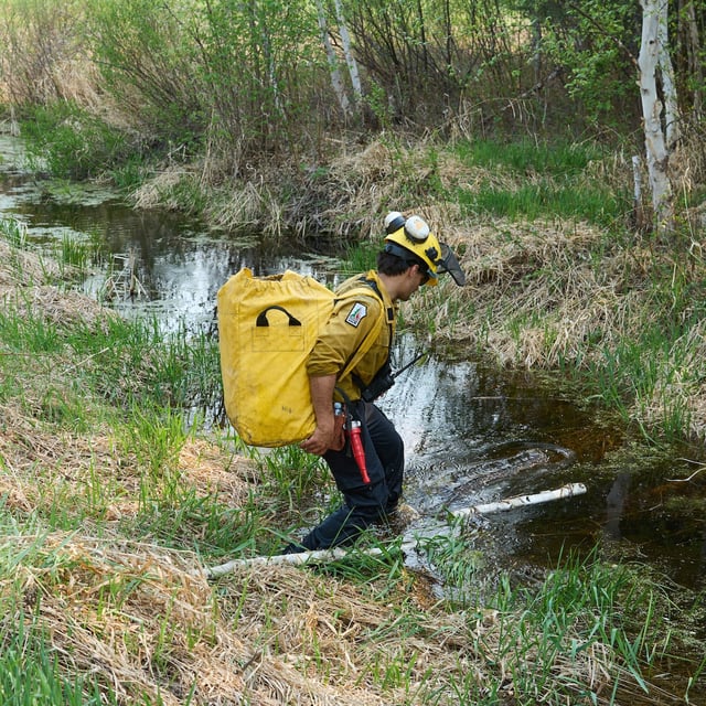 A member of the Manitoba Wildfire Program crosses a stream with a new hose on his back as fire crews continue to fight wildfires around Lac du Bonnet, Man., Thursday, May 15, 2025. THE CANADIAN PRESS/David Lipnowski