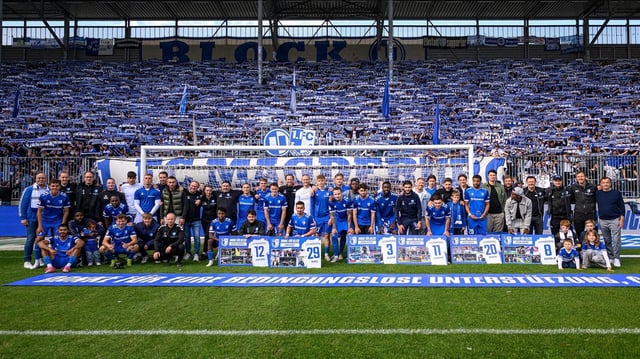 Die Mannschaft des 1. FC Magdeburg steht in Blau vor einer Tribüne voller Fans, ebenfalls in Blau und Weiß.