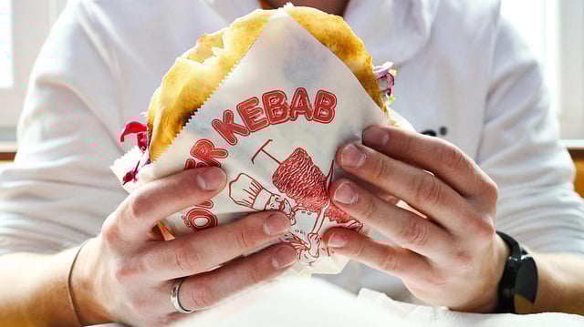 Ein Mann hält einen Döner Kebab in einer bedruckten Dönerverpackung.