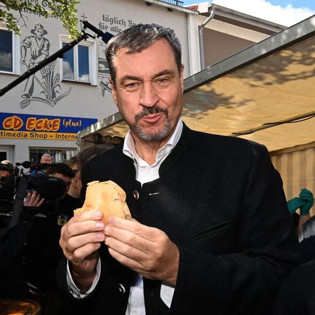 Der bayerische Ministerpräsident Markus Söder (CSU) posiert mit einer Fleischsemmel neben einem Grillmeister beim Schweine-Wettgrillen in Viechtach.