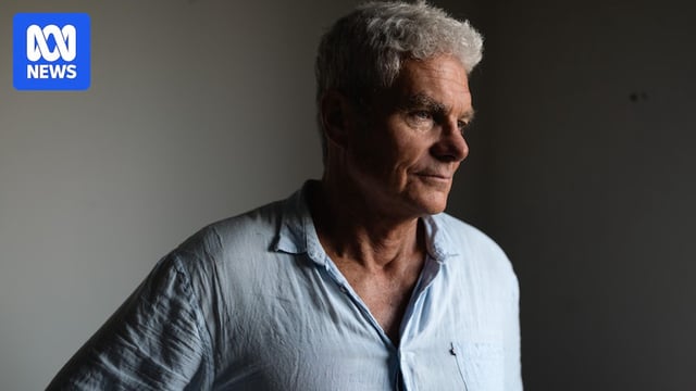 A man standing in a room looks to his side, with a serious and focused expression. Part of his face and the room is in shadow.
