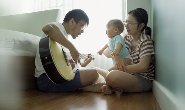 Asian father singing and playing acoustic guitar with his little baby boy and mother on holidays together. Happy family at home.