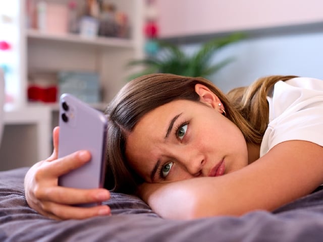 Depressed Teenage Girl Lying On Bed At Home Looking At Mobile Phone
