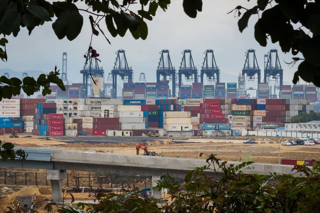 Stacked shipping containers, gantry cranes, and a construction site are seen at the Yantian International Container Terminal through tree branches, with workers and machinery visible in the foreground, on April 12, 2025 in Shenzhen, China.
