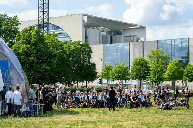 Die knapp 40 Teilnehmenden des "Parlaments der Menschen" sitzen in einer Reihe und hören einem Redner zu, im Hintergrund das Kanzleramt