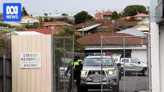 A police officer opens a wire gate, behind which a police vehicle is parked. Two other police vehicles are also visible