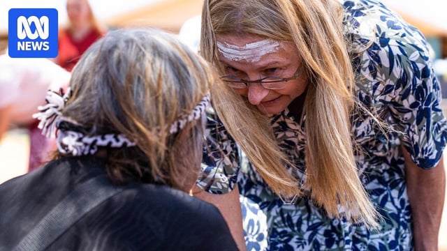 A woman with blonde hair and glasses leans down to talk to an Indigenous person.