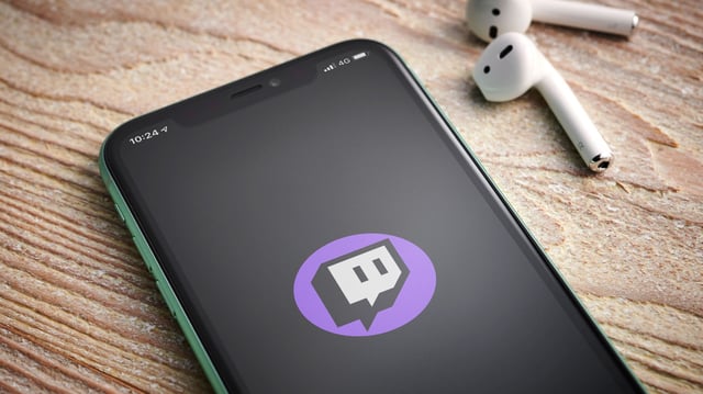 Smartphone mit dem Twitch-Logo auf dem Bildschirm liegt auf einer Holzoberfläche. Daneben befinden sich zwei weiße kabellose Ohrhörer.