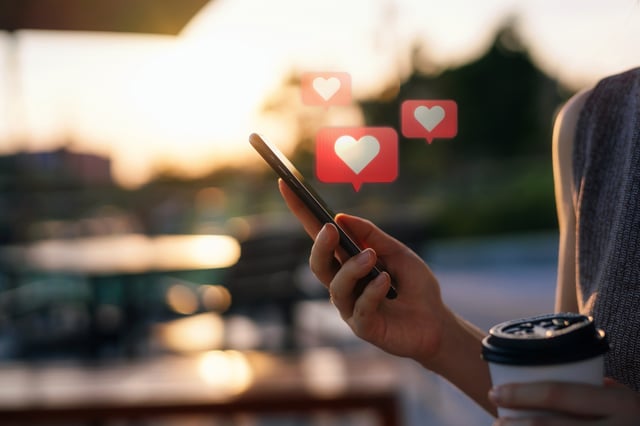 Close up of young woman checking social media network on smartphone on the go in the city. Mobile phone with heart emoji speech bubble message on the screen. People engaging in networking with technology. Social media addiction concept.