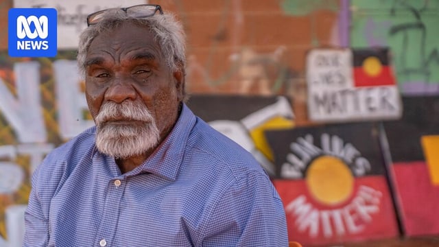 Aboriginal man, gray hair, in a button up shirt sitting in front of a sign that reads 'Blak Lives Matter' on an Aboriginal flag