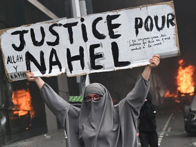TOPSHOT - An attendee holds a banner reading "Justice for Nahel" as cars burn in the street at the end of a commemoration march for a teenage driver shot dead by a policeman, in the Parisian suburb of Nanterre, on June 29, 2023. Violent protests broke out in France in the early hours of June 29, 2023, as anger grows over the police killing of a teenager, with security forces arresting 150 people in the chaos that saw balaclava-clad protesters burning cars and setting off fireworks. Nahel M., 17, was shot in the chest at point-blank range in Nanterre in the morning of June 27, 2023, in an incident that has reignited debate in France about police tactics long criticised by rights groups over the treatment of people in low-income suburbs, particularly ethnic minorities. (Photo by Bertrand GUAY / AFP) (Photo by BERTRAND GUAY/AFP via Getty Images)