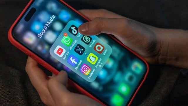 A boy's hands holding an iPhone showing the icons of various social media apps including YouTube, Facebook, X, TikTok, and Instagram.