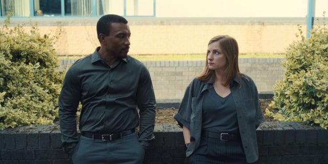 Ashley Walters and Faye Marsay leaning against a brick wall while looking at each other in Adolescence.