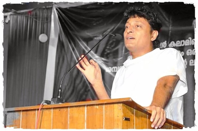 Boby Chemmanur speaking at a podium, wearing a white outfit, gesturing with his right hand during a speech.