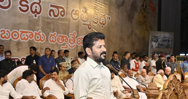 Telangana Chief Minister A. Revanth Reddy speaks at a podium during a public event, surrounded by seated dignitaries and attendees. The background features a large Telugu-language banner and historical imagery. The ornate wooden podium and richly colored floor tiles add to the ceremonial atmosphere.