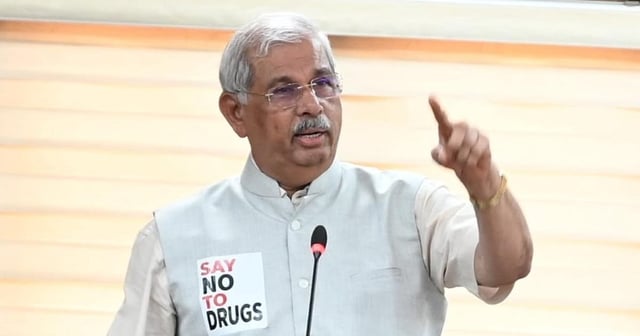 Kerala Governor Rajendra Arlekar is standing at a podium, delivering a speech. He is wearing a beige kurta with a light gray Nehru jacket. A microphone is positioned in front of him, and he is making a gesture with his right hand. On his jacket, there is a badge that reads "SAY NO TO DRUGS." The background features beige blinds, and a security personnel member is partially visible behind him.