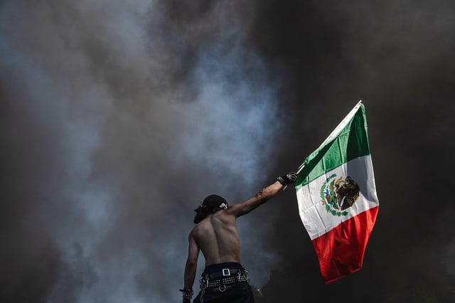 A protester holds up a Mexican flag while standing in front of smoke billowing from burning cars on June 8, 2025 in Downtown Los Angeles, California.