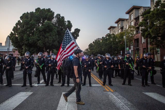A man walks holding an American flag in front of the Los Angeles Metro Police while they try to disperse demontrators at the Little Tokyo neiborhood during protest in response to federal immigration operations in Los Angeles, on June 9, 2025. US President Donald Trump on June 9 ordered active-duty Marines into Los Angeles, vowing those protesting immigration arrests would be “hit harder” than ever. Protests in Los Angeles, home to a large Latino population, broke out on June 6, triggered by immigration raids that resulted in dozens of arrests of what authorities say are illegal migrants and gang members. (Photo by Apu GOMES / AFP) (Photo by APU GOMES/AFP via Getty Images)