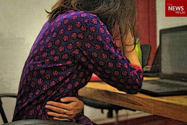 A woman seated at a table, facing a computer, has her arm around her stomach as though in pain. 
