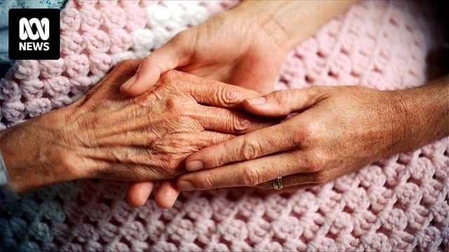 A woman's hand clasps an elderly person's had as it rests on a crocheted blanket.