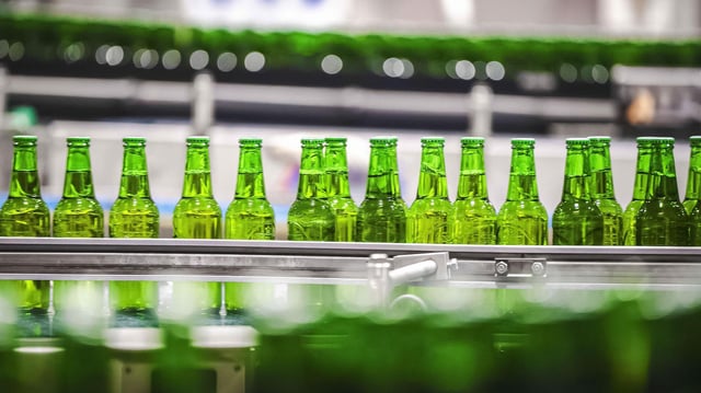 Bierflaschen mit Kronkorken laufen durch die Produktion einer Brauerei über die Förderbänder einer Befüllungsanlage.