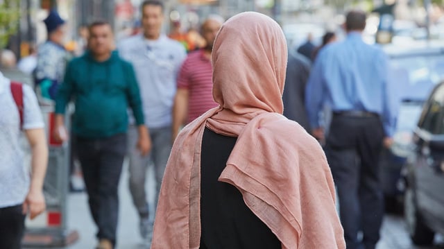Eine Frau mit Kopftuch steht auf einem Bürgersteig.
