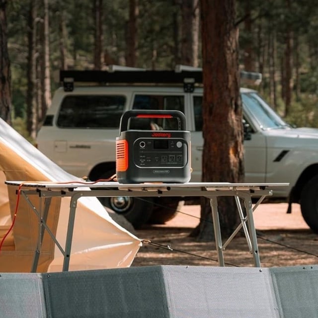 the Jackery explorer 1000 portable power station sits on a camping table in the woods while solar panels charge it and a rugged vehicle is parked behind it