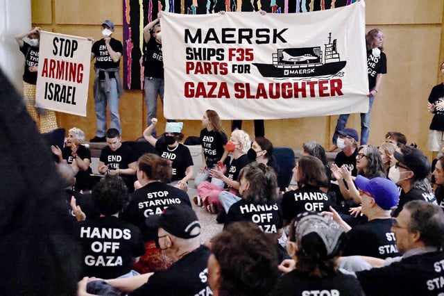 People seated on the ground wearing black shirts with white text that reads "HANDS OFF GAZA", "STOP STARVING GAZA" and "END THE SIEGE" clap and cheer while others hold banners reading "MAERSK SHIPS F35 PARTS FOR GAZA SLAUGHTER" and "STOP ARMING ISRAEL" during an indoor rally
