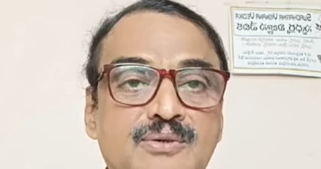 A middle-aged man with short black hair, a trimmed moustache, and large maroon-rimmed glasses is seen speaking. He is wearing a multicolored checkered shirt and is positioned against a plain indoor background with a Telugu signboard visible on the wall behind him.