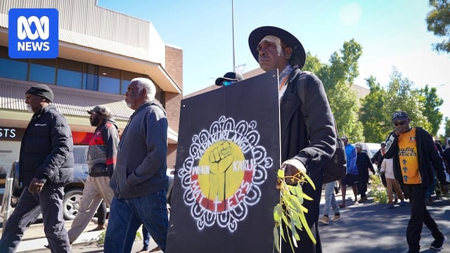 A group of Aboriginal men walk through the town of Alice Springs, one man holding a protest sign. 
