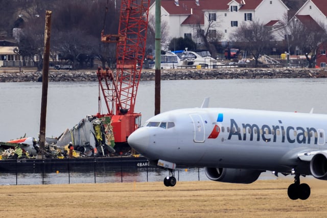 A barge carrying a crane transports parts of the wreckage from the Potomac River as an American Airlines plane lands on the runway, in the aftermath of the collision of American Eagle flight 5342 and a Black Hawk helicopter that crashed into the river, by the Ronald Reagan Washington National Airport, in Arlington, Virginia, U.S., February 5, 2025. REUTERS/Eduardo Munoz
