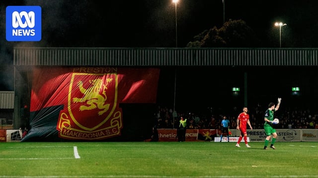 Preston and Heidelberg players on the pitch in front of a huge team flag that says Preston Makedonia F.C