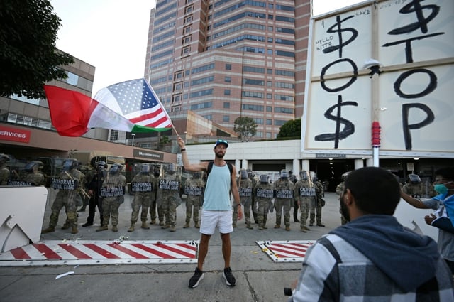 A man stands in front of a row of California National Guard members and waves a combination US and Mexican flag during ongoing protests in response to federal immigration operations in downtown Los Angeles