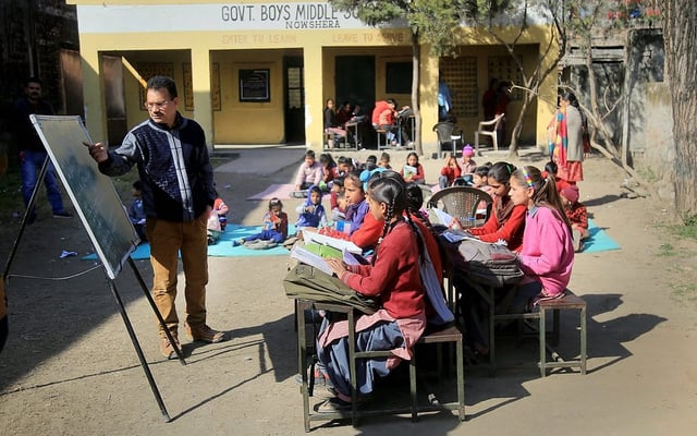 A teacher conducts an outdoor class at a government boys’ middle school in Nowshera, India. Girls in school uniforms sit at desks in the foreground, attentively following the lesson written on a blackboard, while younger students sit cross-legged on mats in the background. The yellow school building and trees surround the open-air learning space on a sunny day.