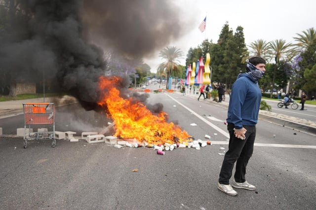 A fire burns as a protester stands across the way from Border Patrol personnel in riot gear and gas masks outside of an industrial park in the Paramount section of Los Angeles on  June 7. (ERIC THAYER/AP)